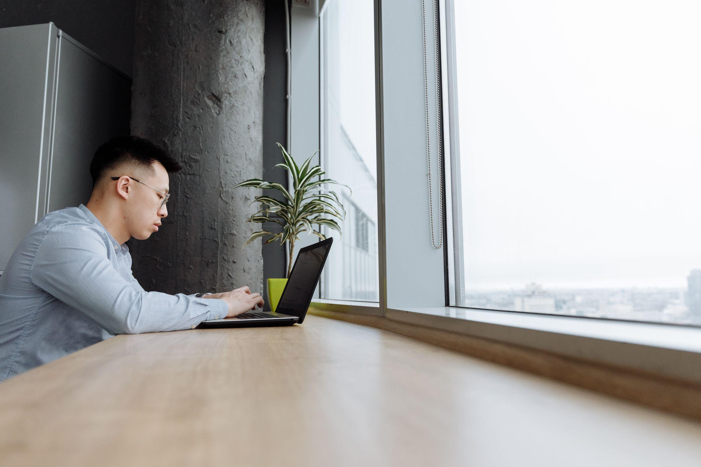 A man working on a laptop at a bright floor-to-ceiling office window, the city faint outside